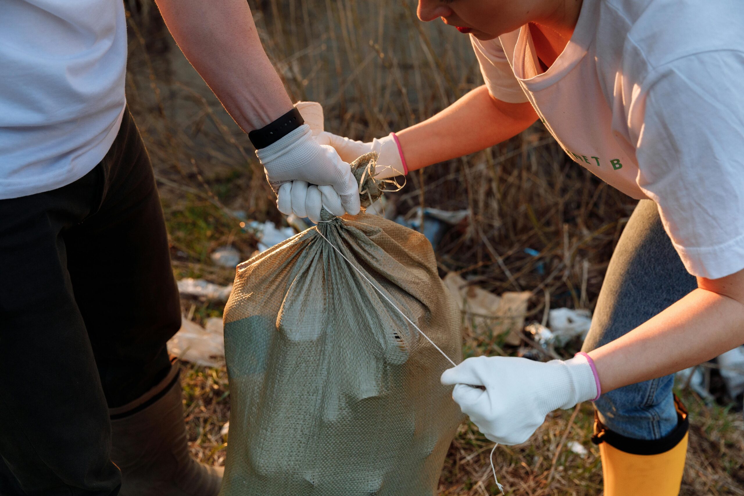 Two people tying a sack of garbage during an outdoor community cleanup activity.